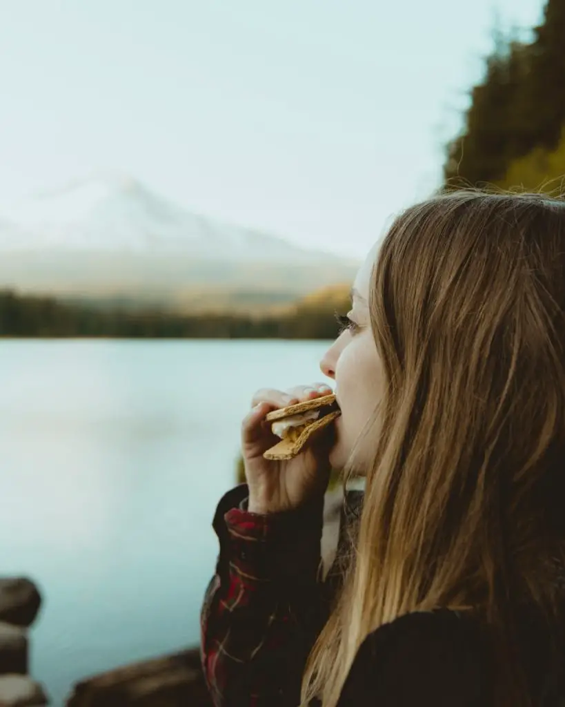 Frau steht und isst Essen mit Blick auf Gewässer und Blick auf den Berg